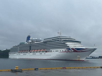 A large white cruise ship, the Arcadia, is docked in the harbor of Bodø, Norway. The ship has a blue smokestack and a prominent Union Jack design on its bow. The sky is overcast and the water is calm. In the foreground, a yellow bollard stands on a wet pier. Other vessels and port infrastructure are visible in the background. The overall scene suggests a cloudy day in a Norwegian port city.