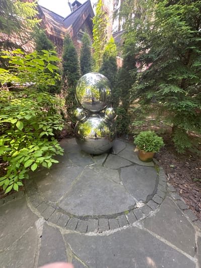 The image shows an outdoor scene in a garden, likely in Gribovo, Russia. The central subjects are a cluster of three large, highly reflective spherical sculptures, appearing to be made of polished metal. These spheres are positioned on a paved pathway made of large, irregularly shaped flagstones bordered by a ring of smaller, rectangular stones.

Surrounding the pathway and sculptures is lush greenery, including tall evergreen trees and bushes with vibrant green leaves. In the background, a portion of a brick building with a visible roofline is present, suggesting the garden is part of a residential property. A small potted plant with dense green foliage sits near the pathway.

The time of day appears to be daytime, with sunlight casting reflections on the sculptures and illuminating the greenery. The weather seems pleasant, conducive to outdoor activities. The scene evokes a sense of tranquility and artistic appreciation, with the sculptures serving as a focal point amidst the natural surroundings. There is no visible text in the image.