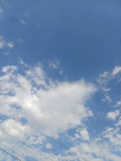 A medium shot captures a bright blue sky filled with scattered white clouds. Several dark power lines are visible diagonally across the lower portion of the frame, with a few small dark birds or insects appearing as specks against the sky. The image is taken from a low angle, looking upward. The location is specified as Jumashuy, Uzbekistan.