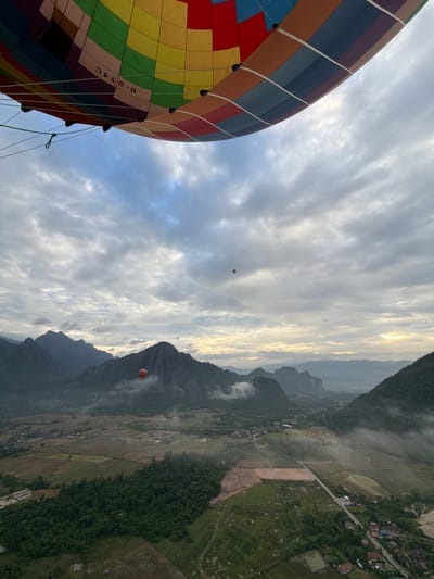 This media file captures a scenic view from a hot air balloon, likely during a sunrise or sunset given the soft, diffused light and the golden glow on the horizon. The scene is set in a mountainous region, possibly in Hong Ngern Village, Laos, as indicated by the presence of lush green hills, terraced fields, and a winding road.

The dominant subject in the foreground is a large, vibrantly colored hot air balloon, its patterned fabric a patchwork of reds, oranges, yellows, blues, and greens. The balloon is tethered and likely in the process of ascent or descent. In the distance, two smaller, red hot air balloons are visible, adding to the sense of wonder and adventure.

The landscape unfolds below, showcasing a rural setting with patches of cultivated land, verdant forests clinging to the slopes of the mountains, and a scattering of houses and buildings that suggest a small village. Wisps of mist or low-lying clouds drift across the valleys, adding a mystical quality to the panorama. The sky is filled with dramatic, textured clouds, some of which are illuminated by the sun's rays, creating a sense of depth and vastness.

The overall mood is one of peace and awe, with the expansive natural beauty and the gentle presence of hot air balloons evoking a feeling of tranquility and escape. There is no visible text other than some lettering on the large balloon, which appears to read "B-U-SAC". The time of day appears to be early morning or late evening due to the quality of light and the presence of clouds.