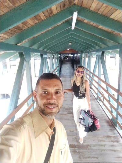 A man takes a selfie while walking along a wooden pedestrian bridge with a woman. The bridge has a green-painted frame and wooden railings. The man wears a tan shirt and a black strap across his shoulder. The woman walks ahead, wearing a black top, white pants, and sunglasses, carrying bags. The bridge's ceiling is made of wooden planks. Lights are attached to the ceiling. The background fades into the distance.