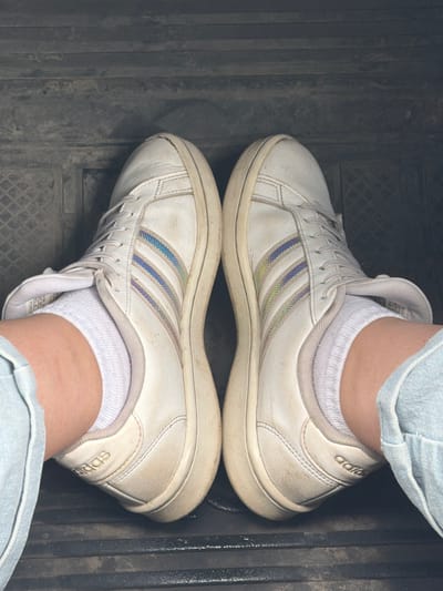 The image shows a close-up, overhead view of a person's feet wearing white Adidas sneakers and white socks. The sneakers have iridescent stripes on the sides and appear worn and slightly dirty. The person is wearing light blue denim jeans. The feet are positioned on a dark, textured surface that resembles the floor mat of a vehicle. The background is blurred and dark. The location is identified as Nezhinka, Russia.