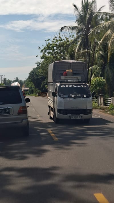 The image shows a white Isuzu truck driving on a road in Padangkasab, Indonesia. The truck has a gray tarp covering its cargo and has several stickers on the windshield, including "BISMILLAH" and "ABANG GEMOT". There is also a silver SUV in front of the truck, with its license plate visible as "B 268 GH". The road is asphalt with a yellow dividing line. Trees and palm trees line the sides of the road, and the sky is partly cloudy. The scene appears to be a sunny day.