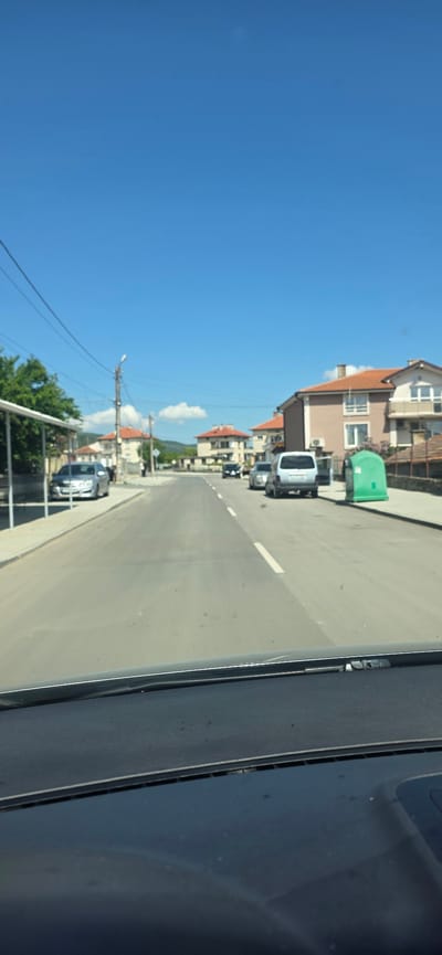 The image depicts a view from inside a vehicle traveling down a road in Chernograd, Bulgaria. The road is asphalt with a single white line dividing the lanes. Buildings line the left side of the road, including a two-story structure with a brown exterior, red tile roof, and a green waste container in front. Several cars are parked along the side of the road, including a silver van. Utility poles with overhead wires are visible against a clear blue sky with scattered clouds in the distance. Vegetation is present near the bottom of the frame, partially obscuring a fence or railing. The right side of the image shows the interior of the vehicle, possibly a window or dashboard.