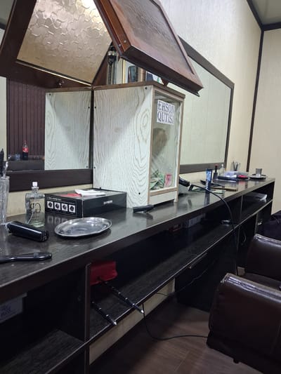 The image shows the interior of a barbershop in Kahramon, Uzbekistan. The scene is set at a wooden counter, reflecting in a large mirror. On the counter, there is a glass display case with a wooden frame, containing cash. A white label on the glass reads "EHSON QUTISI". To the left of the display case, a bottle of clear liquid with a spray nozzle is present, alongside a black electric shaver and a silver oval dish. Behind the bottle, a box labeled "BARBERS" is visible, with a white cloth on top. Further to the right, various barber tools and accessories are scattered, including brushes, scissors, and a cup. A dark-colored barber chair is partially visible in the foreground, on the right side of the image. The walls appear to be paneled with a light-colored material. The overall impression is a functional and moderately organized barbershop.