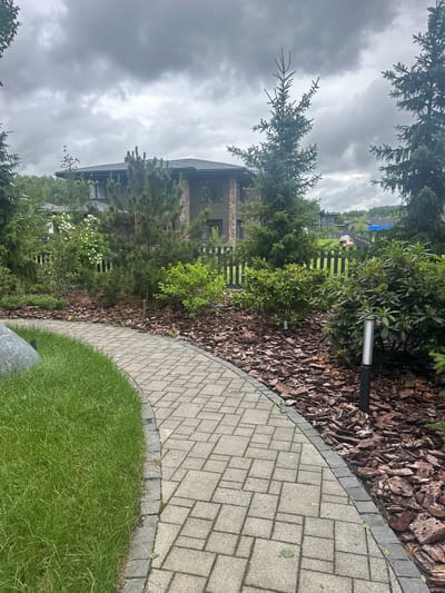 The image depicts a residential property in Uyut, Russia, on an overcast day. The scene features a curved pathway made of light gray pavers, bordered by dark gray stones. To one side of the pathway is a well-maintained lawn of bright green grass. Adjacent to the lawn and pathway is a garden bed mulched with dark brown wood chips. Several evergreen trees, including what appear to be pine trees, are visible in the garden, along with a rhododendron bush. A modern outdoor light fixture stands in the garden bed.

In the background, a multi-story house with a dark roof and a facade combining brick and wood elements is partially visible, surrounded by more greenery. A dark wooden fence runs along the property line, separating the garden from a distant view of other houses and trees. The sky is heavily clouded, suggesting a gloomy or rainy weather condition. No people are visible in the image. The overall impression is one of a landscaped, affluent suburban or semi-rural setting.