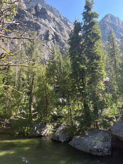 A scenic view of a river flowing through a rocky, forested canyon under a clear blue sky. The foreground features a body of green, clear water with smooth, rounded rocks along the bank. In the background, a steep, rocky mountain face rises sharply, sparsely dotted with evergreen trees and branches. Sunlight illuminates the scene, casting shadows on the rocks and water. The location is not specified as being within the city of Kanawyers, United States, as that is not a known city.