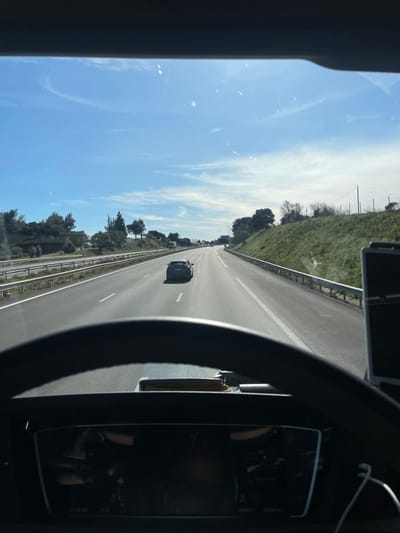 The image presents a view from inside a vehicle, likely a truck, on a multi-lane highway near Tavel, France, on a clear, sunny day. A dark steering wheel and a digital dashboard display, showing "14035 KM" among other indistinct information, are prominent in the lower foreground. Through the windshield, the paved highway extends into the distance, marked with white lines for at least two lanes in each direction. A single dark-colored passenger car, appearing to be a hatchback or small SUV, is positioned in the middle lane, moving away from the viewer. Metal guardrails line both sides of the roadway, separating it from grassy embankments adorned with scattered trees and shrubs. The sky is a bright blue with sparse, wispy white clouds, and direct sunlight causes glare on the windshield. Additional vehicles are faintly visible in the far distance on the highway, and utility poles stand on the right-side embankment further down the road.