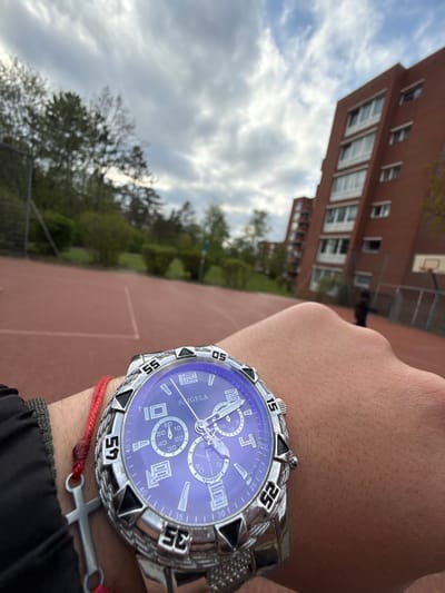 An angled, close-up shot captures a person's left arm, adorned with a large, silver-toned wristwatch and a red string bracelet featuring a silver cross charm. The watch face is a vibrant purple-blue, displaying the brand name "SAIGELA," prominent hour markers, and three subdials. Its chunky bezel is marked with numbers (15, 25, 35, 45, 55) and black triangular inserts.
The background reveals an outdoor setting on a partly cloudy day, suggesting daytime in Lüneburg, Germany. A reddish-brown hard court, possibly a basketball or tennis court, occupies the mid-ground. Beyond the court, a multi-story building with a red facade and numerous rectangular windows stands under the sky. A basketball hoop is partially visible near the building, and green trees dot the landscape, framing the recreational area. The image provides a glimpse of personal style against an urban residential backdrop.