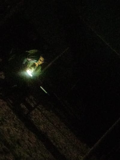 The image displays an outdoor nighttime scene in Al-Mawasi, Palestinian Territories, under conditions of extremely low light. A single male individual is visible in the upper-left quadrant, partially illuminated by a strong, localized white light source positioned below his face. The individual appears to have light-colored hair or a beard and is wearing an upper garment with a green or camouflage pattern. His face is directed towards the upward-shining light source. Below and to the left of the individual, a faint, elongated object emitting a green glow is discernible. The surrounding environment consists of dark, indistinct ground textures, suggestive of dirt or rough terrain, with faint diagonal lines and patterns across the lower half of the frame.
