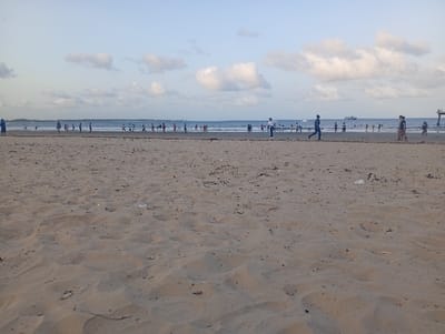 The image displays a sandy beach scene in Shella ward, Kenya, under a light blue sky with scattered white cumulus clouds. The foreground consists of rippled, light brown sand containing scattered dark organic debris and one prominent white plastic item near the center-right. Along the ocean's shallow waterline in the midground, approximately 25-30 individuals are present, engaged in various unidentifiable activities. Three distinct groups of people are closer on the sand: one individual in a blue garment stands on the far left, a male in dark clothing is captured mid-stride running right-to-left with another male standing slightly behind him to his left, and a group of three (two adults, one child) walks towards the far right. The ocean surface contains a larger, green-hulled vessel in the mid-distance to the right and a smaller, white speedboat further left. A pier or dock structure extends into the water from the far right of the frame. A distant landmass is faintly visible on the left horizon.