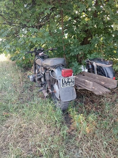 An abandoned, dusty black motorcycle is parked on overgrown grass under a large, leafy tree. The motorcycle has a visible license plate with the numbers "19:45" and Cyrillic letters "ДОЯ" below. The rear wheel of the motorcycle is partially obscured by a stack of weathered wooden planks. In the background, the foliage of the tree creates a dense canopy, with sunlight filtering through. The environment appears rural or on the outskirts of a settlement. The image was captured in the city of Vuhlehirsk, Ukraine.