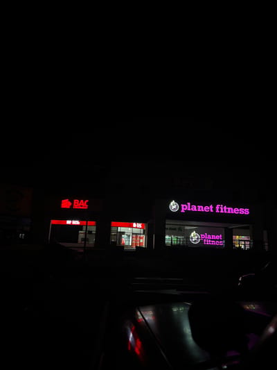 The scene depicts a nighttime view of a commercial establishment located in Nuevo Santiago, Panama, observed from a low-angle perspective, possibly from within a vehicle. The foreground shows the dark, reflective surface of what appears to be a vehicle's dashboard or hood, with red light reflections from the signs ahead.
The primary subjects are two illuminated businesses in a multi-unit commercial building. On the left, a "BAC Credomatic" bank branch is visible. It features a prominent red illuminated sign displaying "BAC CREDO" in white lettering, with "CREDMATO" partially visible below it. Two smaller red signs, also bearing the "BAC" logo, are positioned at ground level. The bank's interior is illuminated, revealing its glass entrance doors and internal lighting.
To the right of the bank, a "Planet Fitness" gym is situated. It is identified by a large, prominent purple illuminated sign with "planet fitness" in white text, accompanied by its stylized "judgment free zone" logo. Below this, a smaller, similarly illuminated "planet fitness" sign and logo are visible at a lower level, possibly marking an entrance or window. The gym's interior also appears to be lit.
The overall environment is dark, indicating a nighttime setting. No individuals or discernible actions are visible within the frame. The buildings' facades are dark, emphasizing the bright, commercial signage.