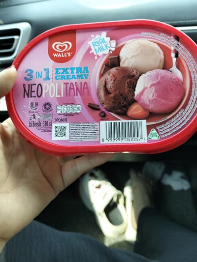 A person's hand is holding a red and pink oval-shaped ice cream container of Wall's Neapolitan flavor in a car. The container has three scoops of ice cream: chocolate, vanilla, and strawberry. The label displays nutritional information and a QR code. The person is wearing a white shoe and dark clothing. The background shows a car's interior. The image does not provide any information about the city of Cotgirek, Indonesia.