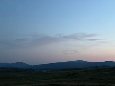 The image depicts a serene, expansive landscape of rolling hills and distant mountains under a pale blue twilight sky. The sky transitions from a lighter blue at the top to a soft pinkish hue near the horizon. Wisps of delicate clouds are scattered across the upper half of the sky.

In the mid-ground, a series of dark, silhouetted mountain ranges recede into the distance, their shapes softened by the fading light. The foreground consists of a vast, dark green grassy field, uneven and textured, with some darker patches suggesting rough terrain or shadows. On the right side of the field, a small herd of dark, indistinct shapes, likely livestock such as cows or horses, are gathered. The overall atmosphere is tranquil and natural, indicative of a rural or wild open space at dusk. The location is specified as Kuseevo, Russia.