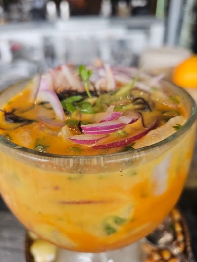 A close-up, vertically oriented image displays a clear glass filled with a vibrant orange-yellow liquid, strongly suggestive of a Peruvian dish like ceviche or leche de tigre. The glass is positioned horizontally across the frame, with its contents sharply in focus. Within the glass, thinly sliced red onions with their distinct purple and white layers are prominently visible, interspersed with fresh green cilantro leaves. Lighter-colored chunks, likely fish or seafood, are submerged in the opaque, richly colored liquid. Darker, thin, curly strands, possibly seaweed or a dark herb, add textural variation.

The background is softly blurred, providing context without distracting from the main subject. To the upper left, a bright orange fruit, possibly a mandarin or small orange, is partially visible. On the right, further in the background, a container holding what appears to be crunchy corn or nuts can be discerned. The scene suggests an indoor setting, possibly a restaurant or home. No specific activity is depicted, but the dish appears ready for consumption. No text is visible in the image. The location context is Homestead, United States of America.
