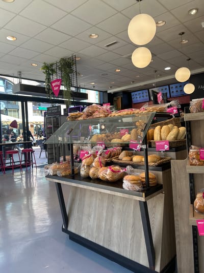 The image shows the interior of a bakery in Groningen, Netherlands. The bakery features a display counter filled with various baked goods, including bread, rolls, and pastries, all packaged in plastic bags. The counter is made of wood and black metal. Above the counter, large spherical pendant lights hang from the ceiling. In the background, through a large glass window, an outdoor seating area with tables and chairs is visible, with people seated and interacting. There are also digital menu boards displayed on the wall behind the counter. Green plants hang from a black metal structure near the ceiling, and pink triangular signs are prominently displayed. The floor is a light gray, reflective surface.