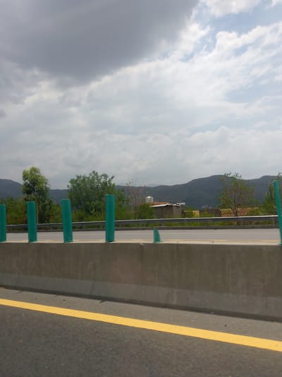 The image shows a roadside view, likely from a moving vehicle, on a cloudy day. In the foreground, a concrete barrier runs horizontally across the frame, with a yellow line painted on the asphalt road in front of it. The barrier is topped with a metal railing, and several green cylindrical markers are positioned behind the barrier. Beyond the barrier, lush green trees and vegetation line a hillside. In the distance, a range of mountains is visible under a heavily clouded sky. The scene appears to be a rural or semi-rural area, possibly a highway or a major road. The image does not contain any visible people or specific identifiable landmarks that would allow for precise geolocation within Abbottabad, Pakistan. However, the general topography of hills and the presence of roads are consistent with areas around Abbottabad.