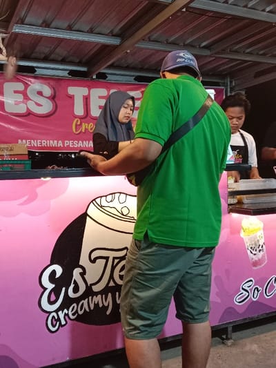 An illuminated food stall operates at night in Sekopek Wetan, Indonesia. A male customer, viewed from behind, wearing a green polo shirt, olive-green shorts, and a blue "RIP CURL" baseball cap, stands leaning on the pink counter of a vendor. He has a black strap from a shoulder bag crossing his back. A young adult female vendor, wearing a grey hijab and a black top, stands behind the counter, looking down as she appears to handle an item or transaction. To her right, another young adult male vendor, wearing a white apron, is partially visible also behind the counter, looking down. The stall features a pink exterior with a "Es Teh creamy" logo depicting a large beverage cup and text in white. A pink banner overhead displays "ES TEH" and "MENERIMA PESANAN" (Receiving Orders) in white lettering. The stall is topped with a metal corrugated roof with exposed metal beams. On the right side of the counter, a clear plastic cup containing a layered, colorful beverage with a yellowish topping is visible, along with other plastic containers. Overhead lighting illuminates the scene.