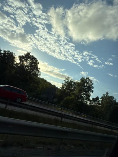 A red station wagon is seen driving on a highway in Kungsbacka, Sweden. The vehicle is in the left lane, moving away from the viewer. The highway is bordered by a guardrail and a dense line of trees and vegetation on the right. The sky above is a clear blue with scattered white, wispy clouds, illuminated by the sun which is partially obscured by the trees. The sun's rays are creating a lens flare effect visible through the foliage. The overall scene suggests daytime travel on a road surrounded by nature.