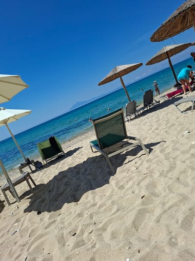 The image depicts a beach scene in Limenaria, Greece. The foreground is dominated by sand with scattered pebbles and debris. Several beach chairs are present, some occupied and some empty. In the middle ground, the turquoise ocean stretches to the horizon, with distant mountains visible. Three thatched umbrellas provide shade, and a few people are seen near the water's edge and in the sea. One person in a blue top and dark shorts is standing on the beach, and another person in dark shorts and a grey top is walking into the water. The sky is a clear, bright blue. The overall impression is a tranquil summer day at the beach.