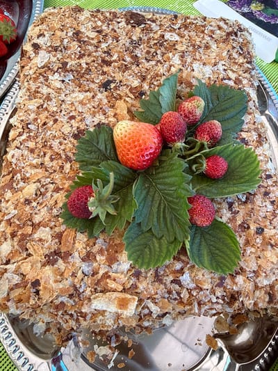 A rectangular cake covered in crumbled pastry and decorated with fresh strawberries and green leaves is presented on a silver platter. The platter rests on a green surface. To the left of the cake, another silver platter holding strawberries is partially visible. A white utensil with "FARBERWARE" printed on it is positioned behind the cake on the right. The overall scene appears to be an outdoor setting, possibly in Anglininkai, Lithuania, indicated by the mention of the city. No people are visible in the image, and no actions or interactions are depicted. The lighting suggests it is daytime.