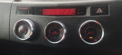 The image shows a close-up of a car's climate control panel. The panel has a digital display showing "OUT SIDE 27°C 12:26". There are three circular knobs below the display. The left knob has symbols for defrosting and fan speed. The middle knob has symbols for airflow direction. The right knob controls the A/C and fan speed, with "LO", "AUTO", "HI", and an A/C indicator light illuminated. To the right of the digital display is a hazard light button with a red triangle symbol. The panel is made of black, textured plastic. There are no people or animals visible in the image. The environment appears to be the interior of a car. There is no information available to determine the location of Покосное, Russia in relation to this image.