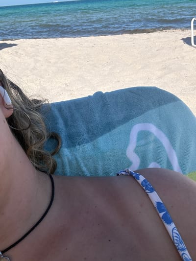 The image displays a close-up, slightly elevated perspective of a person reclining on a beach in Hallandale Beach, United States, under natural sunlight. The visible subject is a fair-skinned individual, presenting the right side of their head and upper torso. Long, wavy, light brown hair is visible on the left, with a white wireless earbud (AirPod) inserted into the right ear. The right shoulder, collarbone area, and upper chest are visible, exhibiting a tanned complexion with several small moles or freckles. A thin black cord necklace with a small, unidentifiable pendant is present around the neck. A white swimsuit strap adorned with a blue floral pattern is visible over the right shoulder. The subject is resting on a multi-tone blue and teal beach towel, featuring a prominent white, stylized 'R' or wave-like emblem. Beyond the towel, light-colored sand extends towards a clear turquoise ocean with calm ripples. A faint white object, likely a boat, is visible on the distant horizon line. A partial white structure, consistent with a beach chair frame or similar object, is visible on the sand at the far right of the frame.