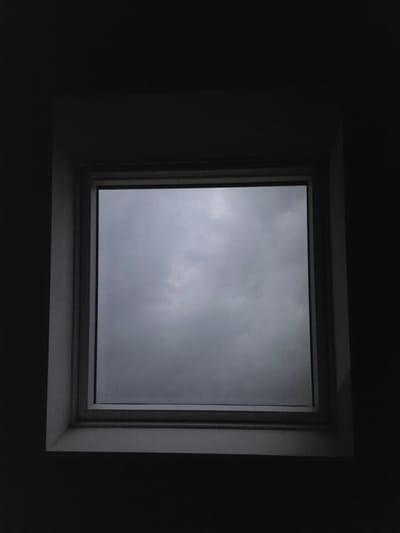 The image shows a square window with a view of a gray, overcast sky. The window is framed by a light-colored material, and the surrounding area is dark, suggesting it is indoors and possibly nighttime or a very dark day. There are no people or specific objects visible outside the window, only the cloudy sky. The scene evokes a sense of gloom or melancholy.

It is not possible to determine the location in the city of Eeklo, Belgium from this image alone.