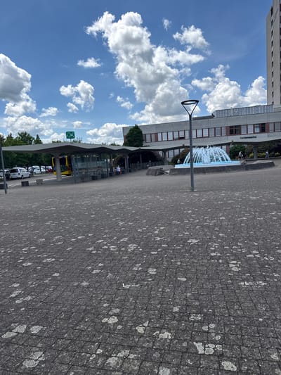 The image shows a street scene in Bottmingen, Switzerland, in front of the Kantonsspital. A fountain is visible. A bus stop is located in the foreground with a bus. Several cars are parked nearby. The sky is blue with cumulus clouds. The pavement is grey with white markings. There are some people nearby at the bus stop.