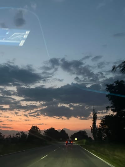 The image depicts a road view at dusk, with a car driving on a two-lane asphalt road. The sky is filled with dramatic clouds, with a gradient of colors from pale blue at the top to orange and pink near the horizon. Silhouetted trees and vegetation line the sides of the road, creating a natural landscape. A tall, slender tree stands out on the right side of the road, while a cluster of trees is visible on the left. The car in front has its taillights illuminated. The scene appears to be captured from inside another vehicle, as suggested by a reflection or glare on the upper left of the image, possibly from a dashboard display. The lighting suggests a low-light environment consistent with twilight or early evening. The image does not contain any specific landmarks or indicators that would definitively place it in Totleben, Bulgaria, but it represents a common roadside scene during sunset in a rural or suburban area.