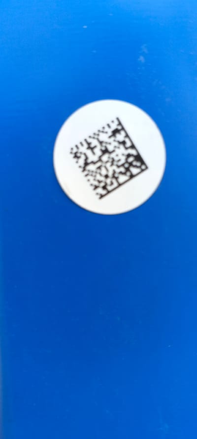 A close-up, overhead shot shows a white circular sticker with a black QR code printed on it. The sticker is positioned on the right side of the frame. The background is a solid, vibrant blue surface, possibly a table or a wall. The lighting is even, with no strong shadows. The image is sharp, with the QR code clearly visible and readable. The overall impression is one of a clean, minimalist presentation.