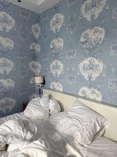 An interior shot of a room in Deauville, France, featuring an unmade bed with white linens. The walls are covered in a light blue and white toile-style wallpaper, displaying recurring ornate patterns of classical or pastoral vignettes framed by decorative scrolls and architectural elements. The bed occupies the foreground, with rumpled white sheets, pillows, and a duvet. A light-colored, upholstered headboard spans the width of the bed. On the left wall, above a dark bedside table, a wall-mounted sconce with a beige cylindrical shade and black metal arm is visible. A clear glass bottle rests on the bedside table. Electrical outlets are present below the sconce. The ceiling is white with decorative molding. No individuals are depicted.