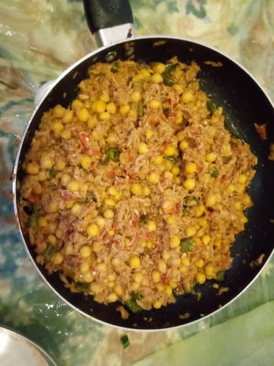 A close-up overhead shot shows a black frying pan filled with a cooked dish. The dish appears to be a mixture of shredded meat or fish, chickpeas, and chopped vegetables such as tomatoes and green peppers. The food is golden brown in color with visible yellow chickpeas scattered throughout. The pan has a silver handle and is placed on a surface that appears to be a patterned tablecloth with green and white swirls. The lighting in the image is overhead, casting soft shadows. No people are visible in the image.
