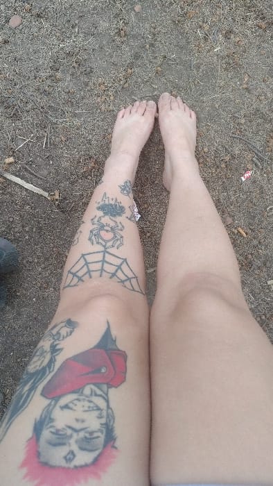 A high-angle, full shot shows a person's bare legs and feet on dirt ground. One leg has multiple tattoos: a spider, a spiderweb, and a grayscale drawing of a person with bright red hair and what appears to be a red garment. The tattoos cover most of the visible portion of that leg. The other leg is bare. The feet are also bare. Some debris litters the ground.