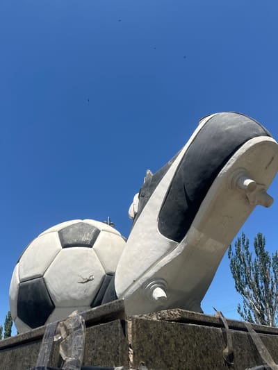 A low-angle, close-up photograph captures a sculpture of a soccer ball and a soccer cleat against a clear blue sky. The soccer ball, rendered in black and white, appears to be made of concrete and shows signs of wear and tear. The soccer cleat, also a concrete sculpture, is positioned next to the ball, with its black and white design prominent. The cleat's studs are visible at the bottom. The sculpture rests on a granite base, which is partially obscured by what appears to be clear plastic sheeting or tape. In the distance, several tall trees with green foliage are visible, along with power lines. A few small, dark shapes, possibly birds or small aircraft, are visible in the sky. The overall scene suggests an outdoor public display, potentially in the city of Lobacheve, Ukraine, given the context provided, though this cannot be definitively confirmed from the image alone.