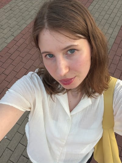In Pocheon-si, South Korea, a young woman with light brown hair and blue eyes takes a selfie outdoors. She is wearing a white collared shirt with subtle texture and a yellow bag is visible over her shoulder. The background consists of a paved walkway with alternating red and gray rectangular stones. The lighting suggests it is daytime, possibly late afternoon given the warm tones.