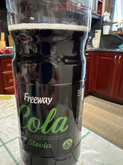 The image shows a close-up of a dark plastic bottle of Freeway Cola with Stevia, lying on its side. The bottle has a black label with green lettering that reads "Cola" and "Stevia" in a stylized font. Below this, in smaller white text, is the slogan "no calories and with sweetener from the plant." The text is in both Greek and English. Some Greek text above the English slogan translates to "without calories and with sweetener from the stevia plant." There are also some Greek words in smaller print near the bottom of the visible label that appear to be related to ingredients or nutritional information.
The bottle is partially filled with a dark, carbonated beverage that has a layer of foam near the opening. Reflections on the bottle suggest the surrounding environment is a kitchen or dining area, with wooden cabinets and possibly a tiled surface visible in the background. The lighting appears to be indoor, suggesting daytime. The overall impression is a product shot focusing on the beverage itself.