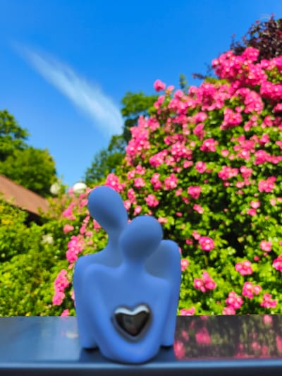 A small, light blue, abstract figurine of two embracing figures with a silver heart on the chest of the front figure is placed on a grey metallic surface. The figurine is in the foreground, with a blurred background of lush green foliage and vibrant pink flowers. In the distance, a red-tiled roof of a building is visible, along with more green trees and a bright blue sky with a wispy white cloud. The setting appears to be a garden or outdoor space, possibly in Georgsmarienhütte, Germany, given the style of the building and the blooming flowers typical of the region during warmer months. The overall impression is one of warmth and tranquility.