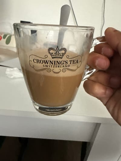 A human hand with light skin tone is holding a clear glass mug by its handle, tilted to the right. The mug contains a light brown, frothy liquid, consistent with a milky beverage like tea with milk or coffee with milk. A white spoon or stirrer is visible inside the mug, partially submerged in the liquid.
The mug features a prominent black logo and text. A crown symbol is visible, and the text reads "CROWNING'S TEA SWITZERLAND," indicating the brand and origin.
The scene appears to be an indoor setting, possibly in Lausen, Switzerland, given the location context and the mug's branding. The background consists primarily of plain white walls or surfaces, with a thin, light-colored wire or cord running horizontally across the upper left. To the right, a darker, rectangular opening or doorway is partially visible. Below the mug, a glimpse of another object with green and red patterns can be seen. The image is taken from a slightly downward angle, focusing on the hand and mug.