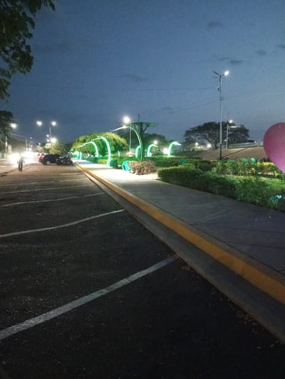 The image depicts an outdoor scene in Villa de Todos los Santos de Calabozo, Venezuela, at dusk or night. A parking lot with white lane markings is in the foreground, and a sidewalk runs alongside it, separated by a curb with a yellow stripe. On the other side of the sidewalk is a landscaped area with hedges, plants, and decorative green-lit arches. Streetlights illuminate the area. In the background, there are trees, and on the far right, a large pink balloon is visible. A person is walking on the side of the road in the middle-left of the image, near several parked cars.