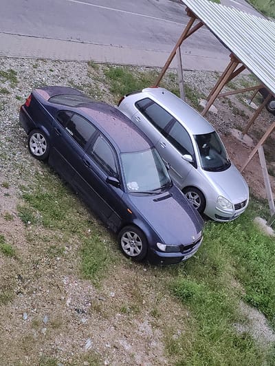 In Krushev Dol, Bulgaria, two cars are parked on a gravel and grass area next to a road. A dark blue sedan is in the foreground, with its front end facing towards the right of the frame. To its right and slightly behind is a silver hatchback. Both cars appear to be wet, possibly from recent rain. In the background, a makeshift wooden shelter with a corrugated metal roof is visible. The surrounding environment consists of a paved road, gravel, and patchy grass. No people are present in the image.