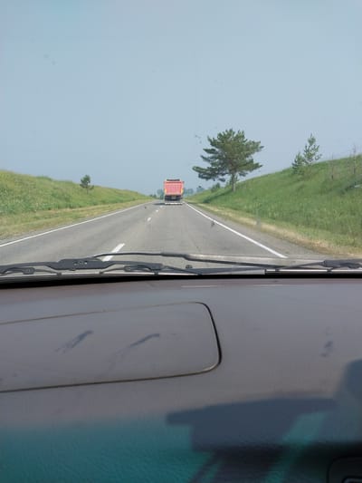 The image depicts a view from inside a vehicle looking out through the windshield at a highway. A red dump truck is visible in the distance on the road. The highway is flanked by grassy slopes with scattered trees. The sky is clear and light blue, suggesting a daytime scene. The foreground shows the interior of the vehicle, including the dashboard and windshield wipers. The location is identified as Novotyryshkino, Russia.