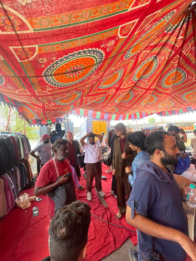 The image shows a gathering of people under a large, red, patterned canopy, likely in a market or public space. The location is Anajpur, India.
People:
- A group of men of varying ages are visible.
- They are standing and sitting on a red fabric or covering.
- Some appear to be looking at something or interacting with each other.
- Some are wearing casual clothing such as t-shirts, pants, and jeans.
Objects:
- A large, red canopy with a circular patterned design covers the area.
- Water bottles are visible.
- A collection of clothing items is stacked near the bottom of the frame.
- A small number of electronic devices are in use
Actions:
- People are standing and sitting, some talking to each other
- Some appear to be taking a break or resting.
Environment:
- The scene appears to be an outdoor location, possibly a market or a temporary setup.
- There are glimpses of buildings and a vehicle in the background.
- The overall impression is that the gathering is part of a market, event, or protest.