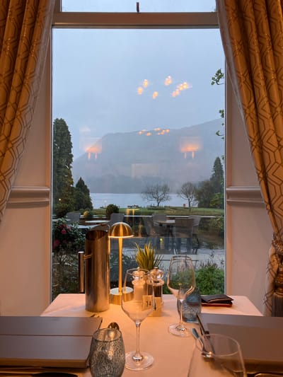 The image shows a view from inside a room in Glenridding, United Kingdom. Through a window framed by patterned curtains, a grey, overcast day is visible over a lake. Trees and buildings are reflected in the window glass. On the table, set for a meal, are wine glasses, a silver ice bucket, a small potted plant, a table number card reading "14", and silverware. The table is covered with a white cloth. The focus is divided between the interior table setting and the exterior landscape.