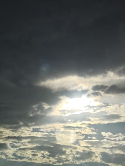 The image is a photograph of a dramatic sky filled with dark, heavy clouds interspersed with patches of bright sunlight. The sun is visible through a break in the clouds, casting a bright glow and creating rays of light that illuminate the surrounding cloud formations. The clouds appear layered, with some darker, denser clouds towards the top of the frame and lighter, more scattered clouds below. The lighting suggests it is either sunrise or sunset, with the low angle of the sun creating a dramatic contrast between light and shadow. There are no people or objects visible in the frame, only the sky and clouds. The overall mood is one of powerful, natural beauty, perhaps even a bit ominous due to the dark clouds. The location context provided is Gricignano di Aversa, Italy.