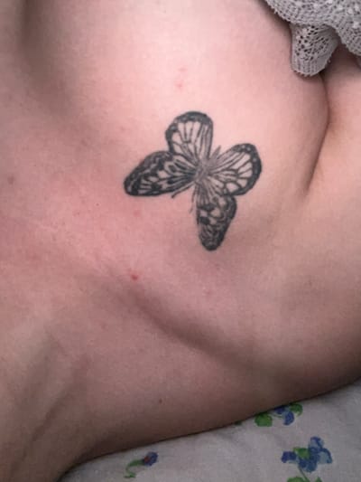 The image shows a black and grey tattoo of a butterfly on a person's skin, likely on the upper chest or shoulder area, given the curvature of the skin. The skin appears to have some pinkish undertones and minor blemishes. A textured grey fabric is visible in the upper right corner of the image, partially obscuring the skin. The lower part of the image shows a white surface with a floral pattern in blue and green. There is no indication of the city of Gorodets, Russia, or any specific urban environment.