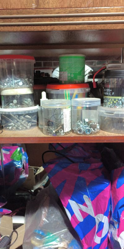 The image shows a close-up, eye-level view of a wooden shelf filled with various hardware items and packaging. On the top shelf, a green cylindrical container with text and an ERC logo is visible. Below this, to the left, are two stacked clear plastic containers filled with screws and nuts. To the right of these, another clear plastic container with a red lid holds assorted fasteners. Further to the right, a black and silver cylindrical container labeled "Profi" contains nuts and is connected to a red wire.
The shelf below is cluttered with various plastic bags and boxes. A large bag with a pink and blue striped pattern is prominent, as are other plastic bags containing items. The lighting appears to be artificial and is casting shadows, suggesting an indoor environment. The overall scene is one of storage and organization of hardware and related materials. The location is identified as Esipovo, Russia.