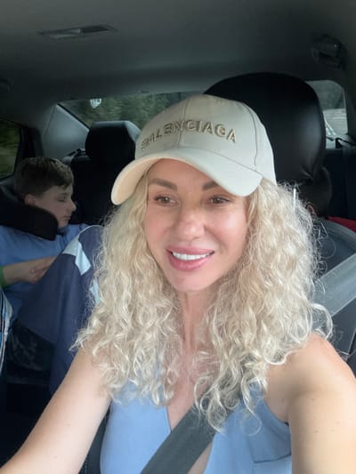 This media file is a selfie taken from inside a car, with the photographer in the foreground and a young boy in the background. The photographer, a woman with blonde, curly hair, is wearing a light-colored baseball cap with visible text that appears to say "BALENCIAGA". She is also wearing a light blue garment. Her expression is happy, with a wide smile and bright eyes. The boy in the background appears to be asleep or resting, leaning against a dark headrest. The setting is the interior of a car, suggesting a journey. The image appears to be taken during the daytime, as natural light filters into the car, but the specific time of day and weather conditions are not discernible. There are no other specific location cues within the image itself that would suggest Navaginskoye, Russia. The overall mood conveyed is relaxed and cheerful, perhaps during a road trip with family.