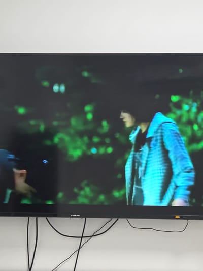 A wide, dark television screen displays a blurred image of two individuals against a background of out-of-focus green lights. On the left, a person wearing a dark helmet or hat is partially visible, with a hand extended. To the right, a person in a light blue, checkered jacket is shown in profile, looking downwards and to the left. The television itself is mounted on a white wall, with black cables hanging loosely below it. A logo reading "NIKAI" is visible on the bottom edge of the screen. The scene appears to be indoors, with the television screen being the primary focus. There is no discernible information within the image to determine the location in Dibba Province, Oman.